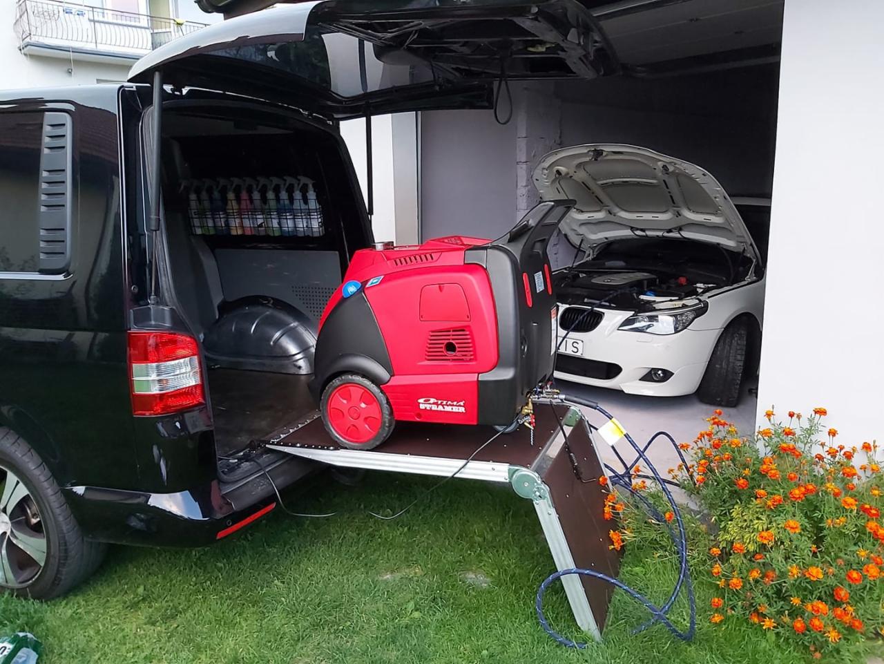 Przenośna, czerwona myjka parowa Optima Steamer umieszczona na wysuwanej platformie z bagażnika czarnego vana, z otwartą maską białego BMW w tle i kwiatami nagietka z przodu.