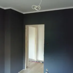 Pomieszczenie po odświeżeniu z ciemnymi ścianami i białym sufitem, widoczne wystające kable od lampy sufitowej oraz fragment podłogi z listwą przypodłogową w przejściu do kolejnego, jaśniejszego...