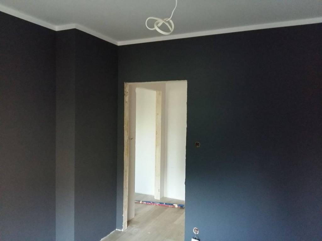 Pomieszczenie po odświeżeniu z ciemnymi ścianami i białym sufitem, widoczne wystające kable od lampy sufitowej oraz fragment podłogi z listwą przypodłogową w przejściu do kolejnego, jaśniejszego...