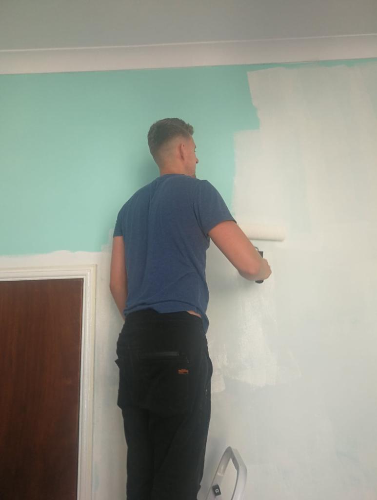 Mężczyzna w niebieskiej koszulce maluje ścianę wałkiem na biało, stojąc na drabinie. Widoczny fragment pomalowanej ściany w kolorze miętowym, drzwi z brązową ościeżnicą w tle.