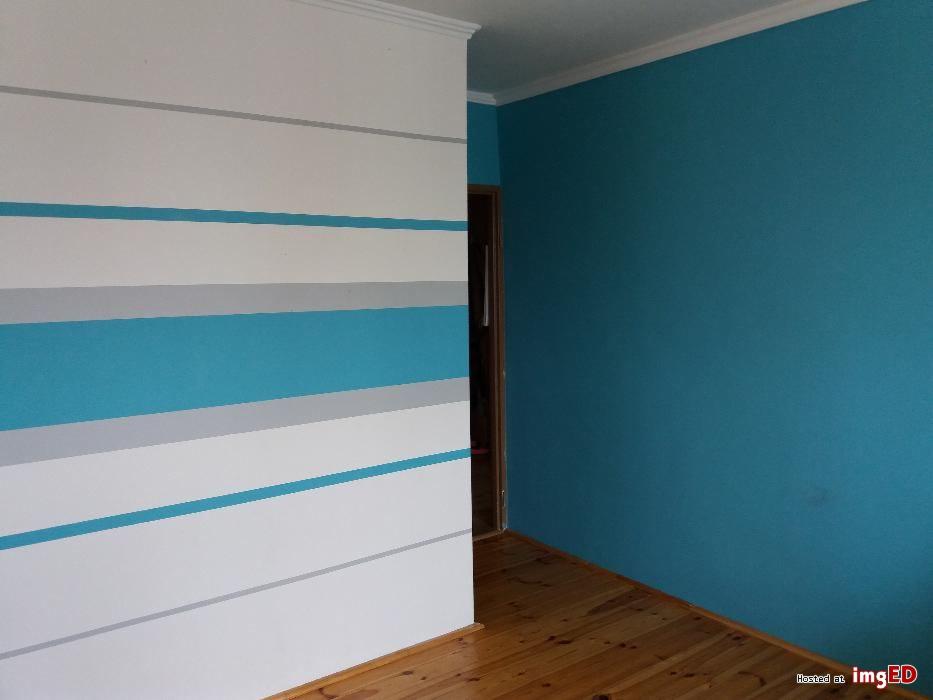 Ściana pomalowana w poziome pasy w kolorach białym, szarym i turkusowym, kontrastująca z jednolitą turkusową ścianą i drewnianą podłogą.