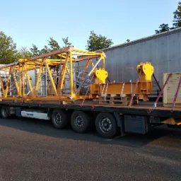 Ciężarówka z naczepą przewozi elementy konstrukcyjne pomalowane na żółto, zabezpieczone pasami transportowymi, na tle błękitnego nieba i zielonych drzew.