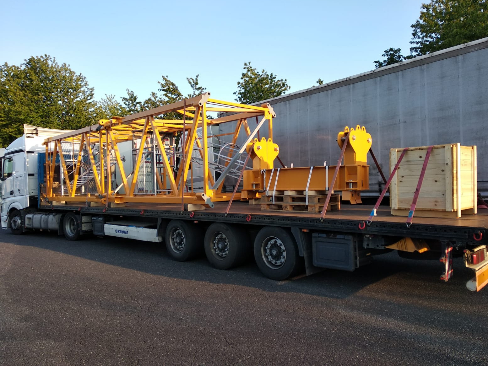 Ciężarówka z naczepą przewozi elementy konstrukcyjne pomalowane na żółto, zabezpieczone pasami transportowymi, na tle błękitnego nieba i zielonych drzew.