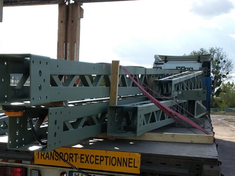 Ładunek ponadgabarytowy w postaci metalowych konstrukcji kratownicowych na naczepie samochodu ciężarowego, zabezpieczony pasami transportowymi, z widocznym znakiem ostrzegawczym 'Transport...