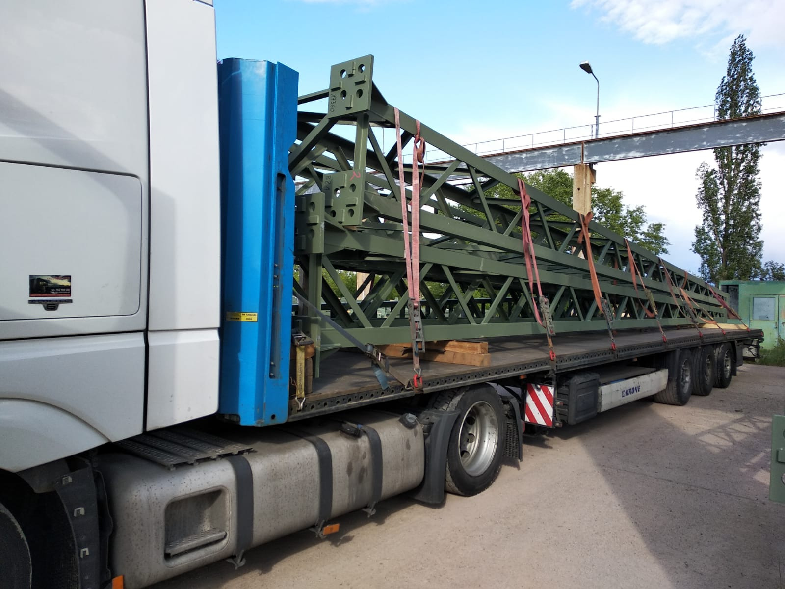 Ciężarówka z naczepą przewożąca elementy konstrukcji stalowej, zabezpieczone pasami transportowymi, widoczne drewniane podkładki.