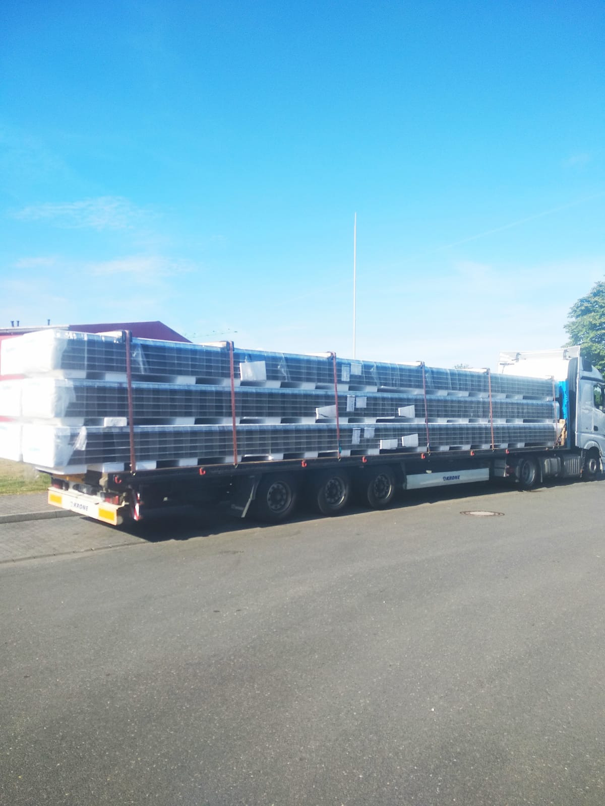 Ciężarówka z naczepą przewozi zabezpieczony ładunek w postaci długich, czarnych elementów, prawdopodobnie okien lub paneli, owiniętych folią, na tle błękitnego nieba i budynków.