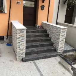 Schody wejściowe do domu wyłożone ciemnymi płytkami i obłożone kamieniem dekoracyjnym, z widocznymi elementami wykończeniowymi i narzędziami pracy.