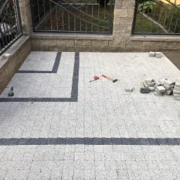 Fragment układanej nawierzchni z jasnoszarej kostki brukowej z ciemnoszarymi pasami, widoczne narzędzia: młotek i miarka, oraz stosy kostki przygotowanej do ułożenia.