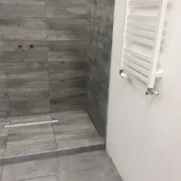 Wykończona kabina prysznicowa z drewnopodobnymi płytkami, widoczny odpływ liniowy i grzejnik drabinkowy na białej ścianie. Minimalistyczny design.