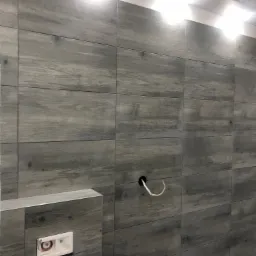 Ściana łazienki wyłożona szarymi płytkami imitującymi drewno, widoczne przyłącza wodne i otwór z kablem elektrycznym, zabudowa stelaża podtynkowego WC z zamontowanym modułem spłukującym.