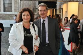 Portret kobiety w białej kurtce i mężczyzny w garniturze, oboje uśmiechnięci, na tle wnętrza z przeszklonymi drzwiami i innymi osobami.