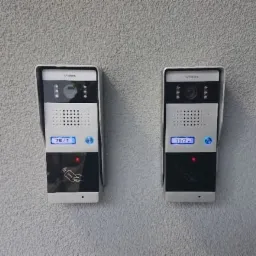 Dwa nowoczesne panele domofonowe z numerami mieszkań (7B/1 i 7B/2) zamontowane na szarej, chropowatej ścianie zewnętrznej.