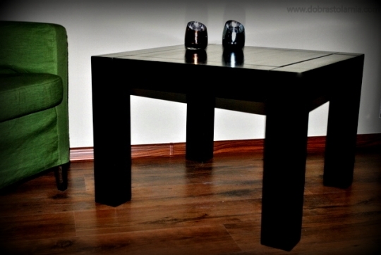 Ciemny, kwadratowy stolik kawowy z dwiema szklanymi świecznikami na blacie, stojący na drewnianej podłodze, obok zielonego fotela.