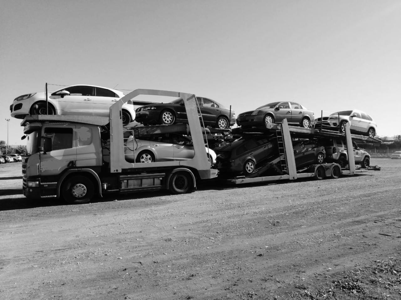 Czarno-białe zdjęcie lawety z załadowanymi samochodami osobowymi na placu, widok z boku, zarys drzew w tle.