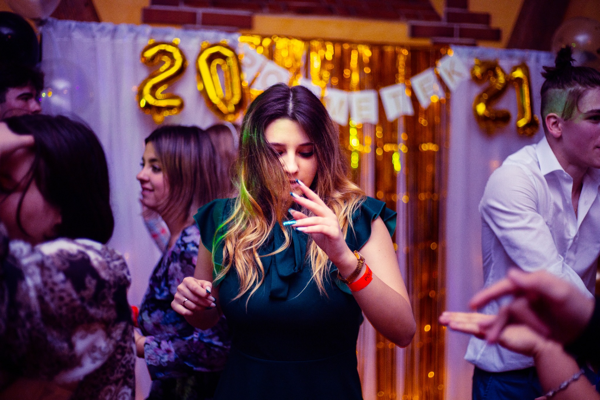 Młoda kobieta w zielonej sukience na tle dekoracji urodzinowych z balonami w kształcie liczby 20 i 21, otoczona innymi osobami.