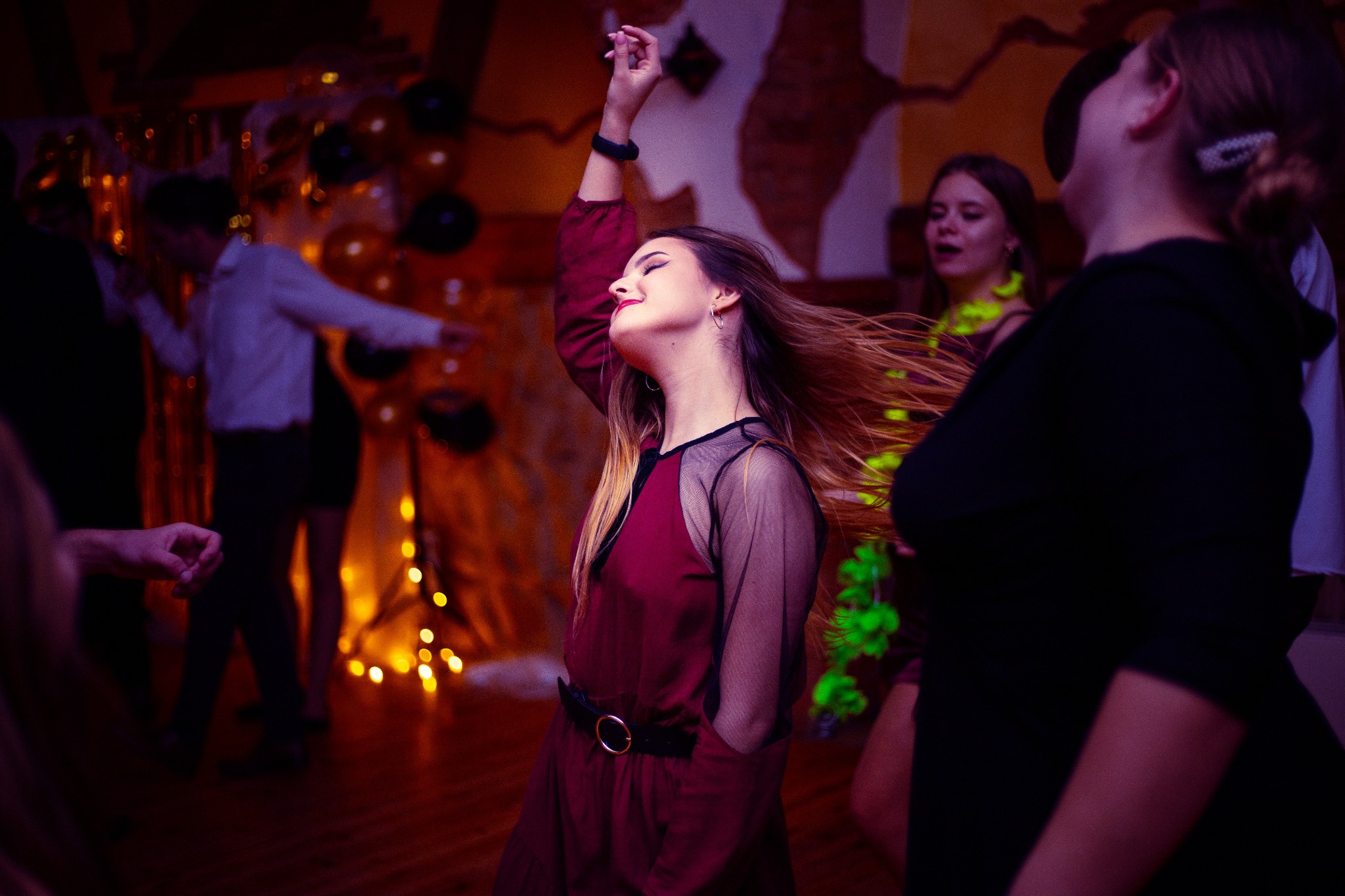 Kobieta w bordowej sukience z siatkowymi rękawami tańczy z uniesioną ręką na parkiecie podczas imprezy, w tle balony i dekoracje świetlne.