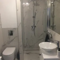 Nowoczesna łazienka z marmurowymi płytkami, kabiną prysznicową typu walk-in, podwieszaną toaletą i umywalką nablatową, minimalistyczna armatura.