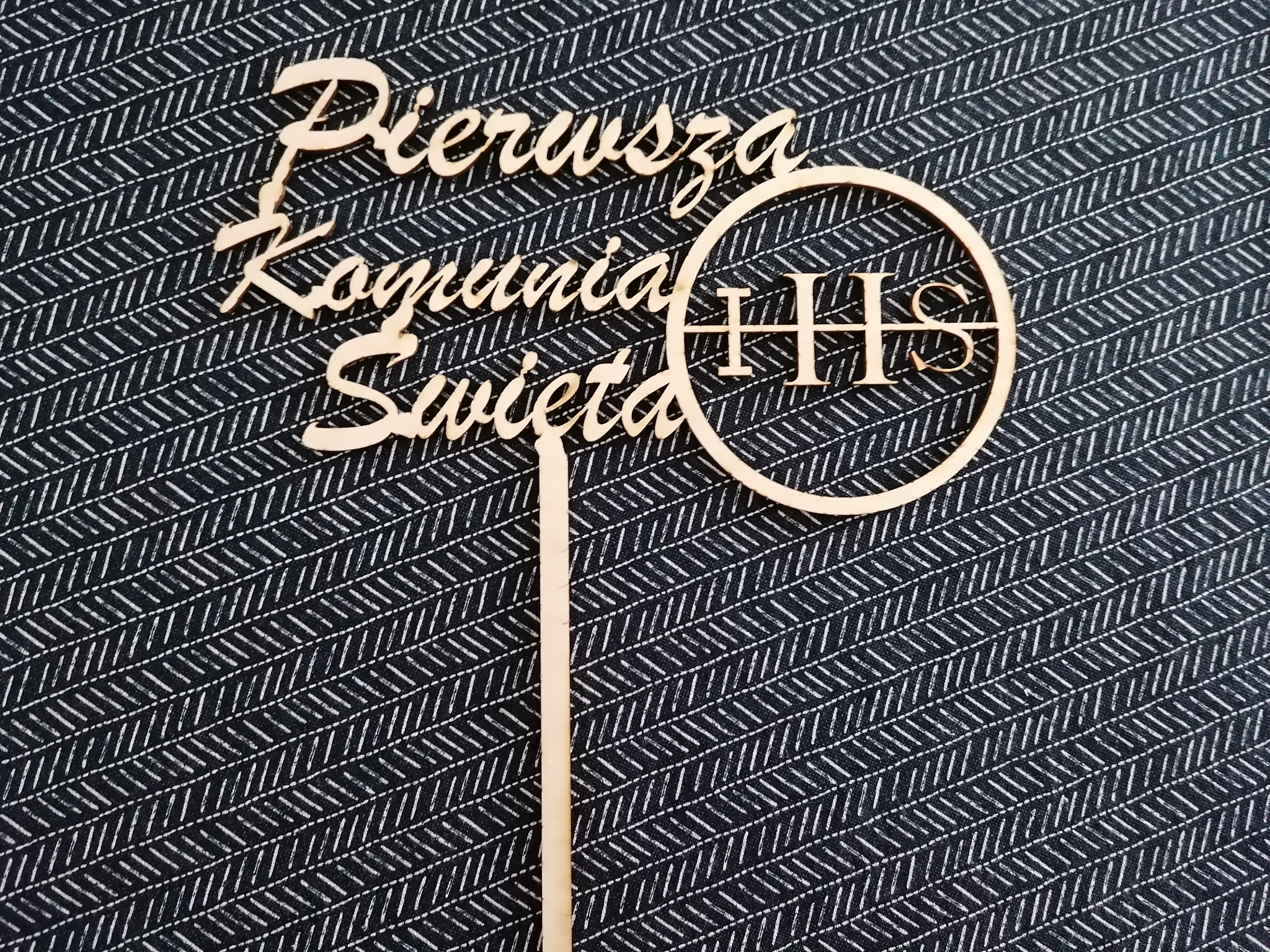 Drewniany topper na tort z napisem 'Pierwsza Komunia Święta' i symbolem IHS na tle ciemnej tkaniny w jodełkę.