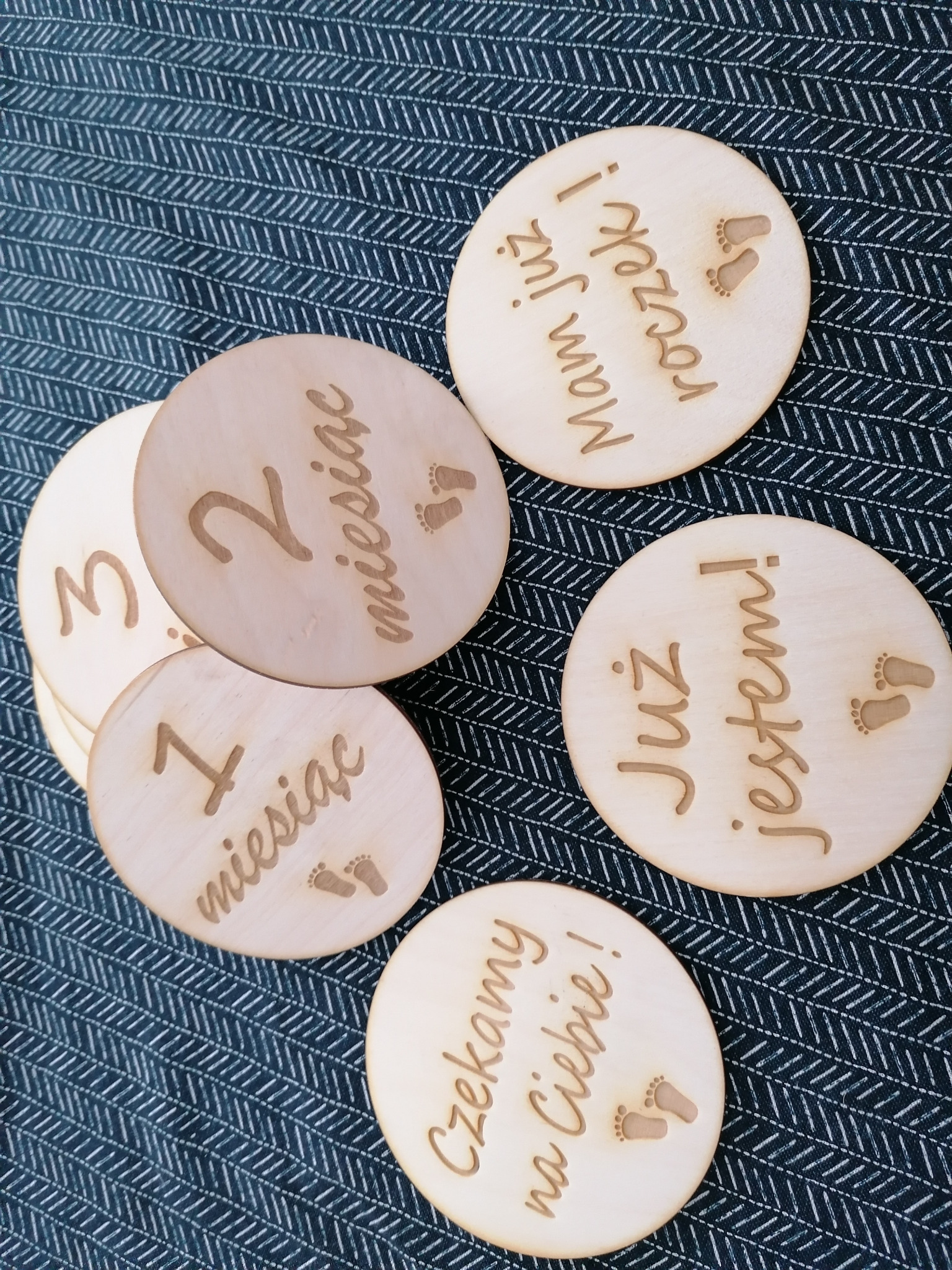 Drewniane krążki z wygrawerowanymi napisami i symbolami stóp, oznaczające kolejne miesiące życia dziecka, ułożone na wzorzystym tle.