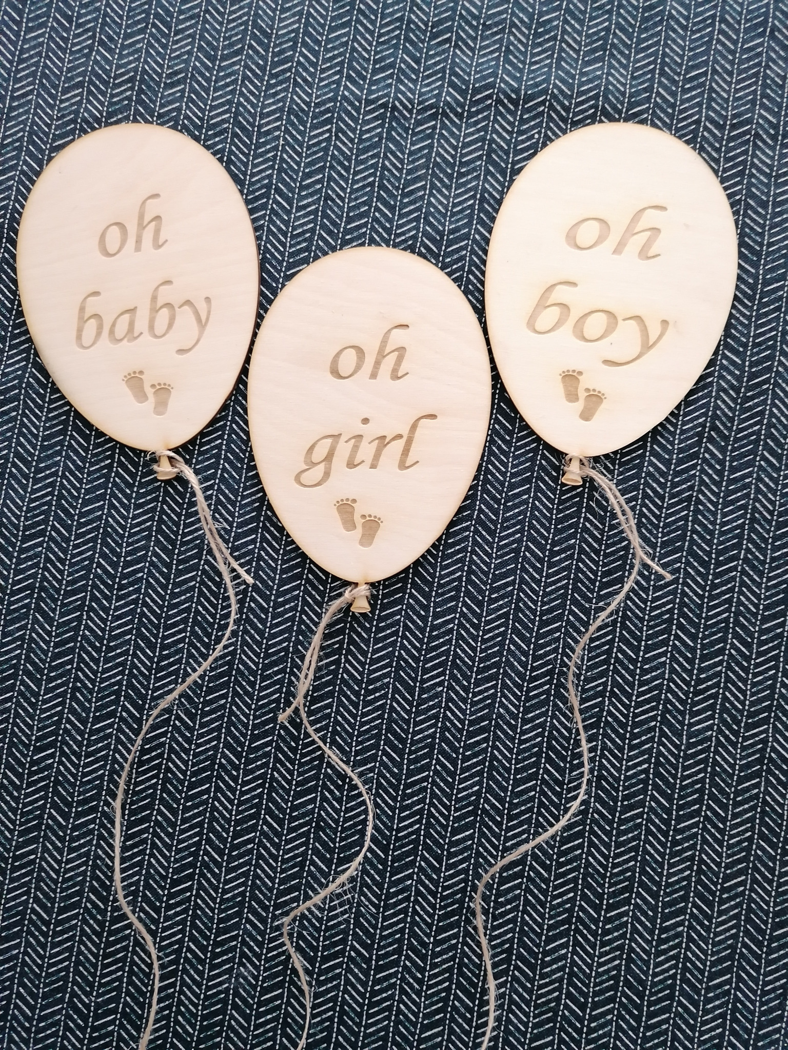 Trzy drewniane balony z napisami 'oh baby', 'oh girl', 'oh boy' i motywem stóp dziecka, leżące na ciemnym materiale w białe zygzaki.