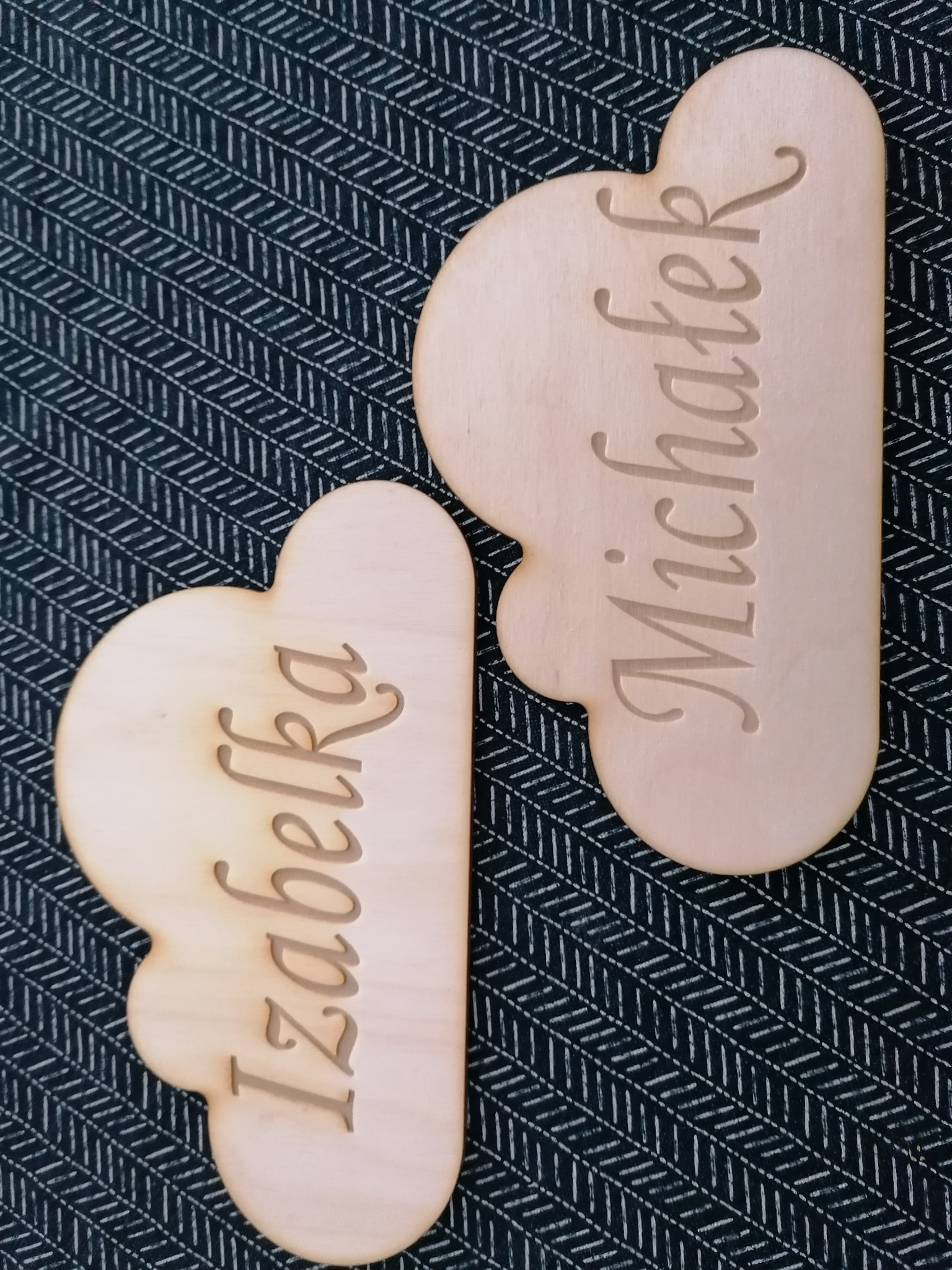 Dwa drewniane, wycięte laserowo zawieszki w kształcie chmurek z imionami Izabelka i Michałek, leżące na ciemnym tle z geometrycznym wzorem.