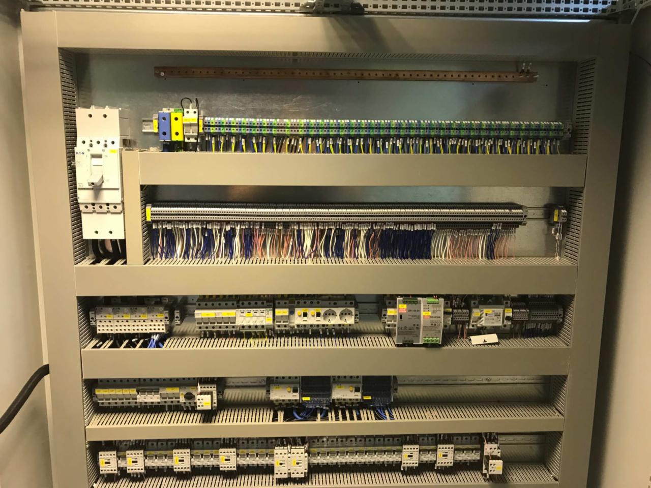 Rozdzielnica elektryczna z widocznymi bezpiecznikami, przewodami i listwami zaciskowymi, uporządkowana w szarym panelu.