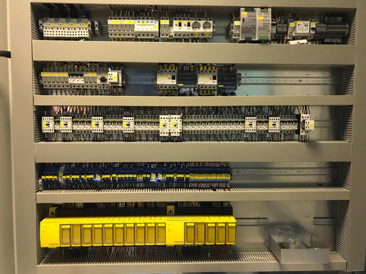 Wewnętrzny widok rozdzielnicy elektrycznej z modułami zabezpieczeń, bezpiecznikami i okablowaniem, uporządkowane w rzędach na metalowych szynach.