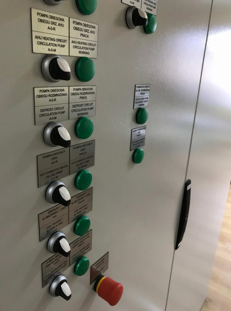 Szara szafa sterownicza z przyciskami i opisami funkcji pomp obiegowych, grzałek i obiegów chłodzenia, widoczny czerwony przycisk awaryjnego wyłączenia.