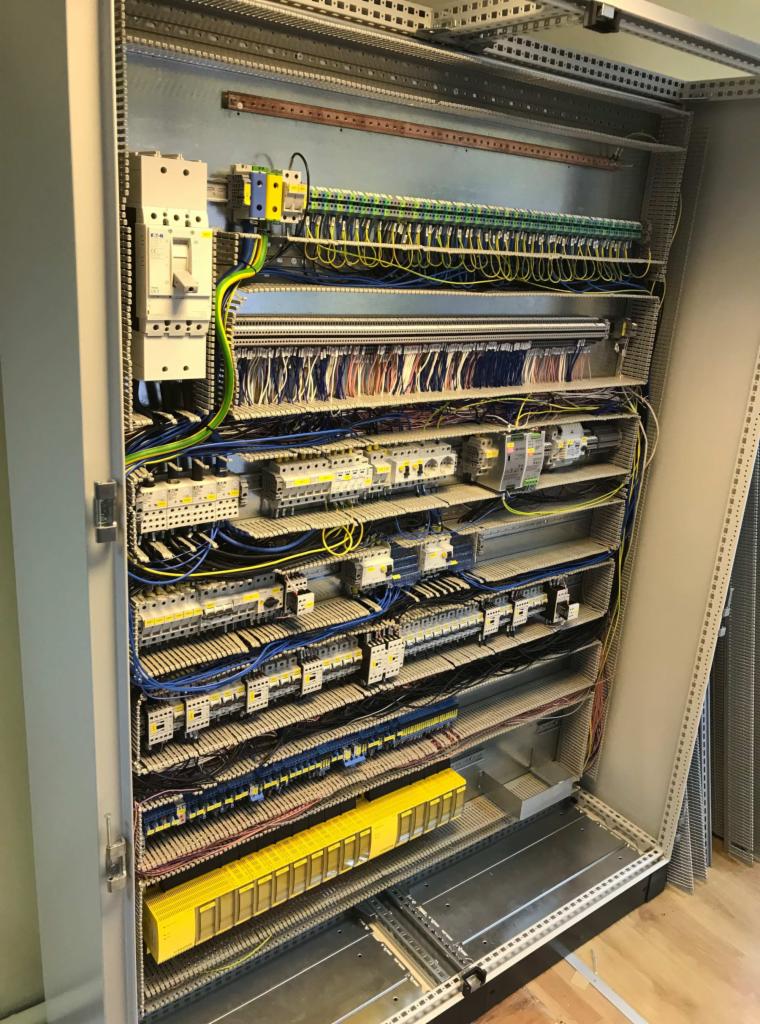 Otwarta szafa sterownicza z widocznymi zabezpieczeniami, przewodami i modułami, uporządkowana instalacja elektryczna wewnątrz metalowej obudowy z perforowanymi ściankami.