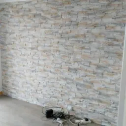 Ściana wykończona panelami imitującymi kamień w odcieniach beżu i szarości, z widocznymi kablami i urządzeniami elektronicznymi leżącymi na podłodze z paneli w kolorze jasnego drewna.