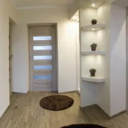 Jasne wnętrze przedpokoju z drewnopodobną podłogą, dwiema parami drzwi, w tym jedne z przeszkleniami, oraz wnęką z trzema półkami oświetlonymi punktowo, na których stoją małe doniczki z roślinami.