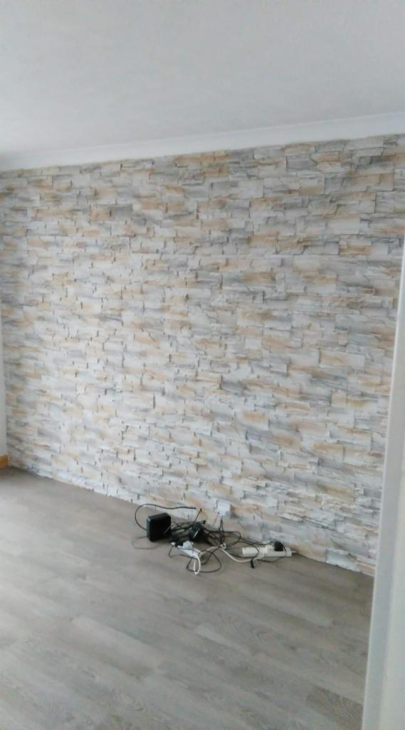 Ściana wykończona panelami imitującymi kamień w odcieniach beżu i szarości, z widocznymi kablami i urządzeniami elektronicznymi leżącymi na podłodze z paneli w kolorze jasnego drewna.