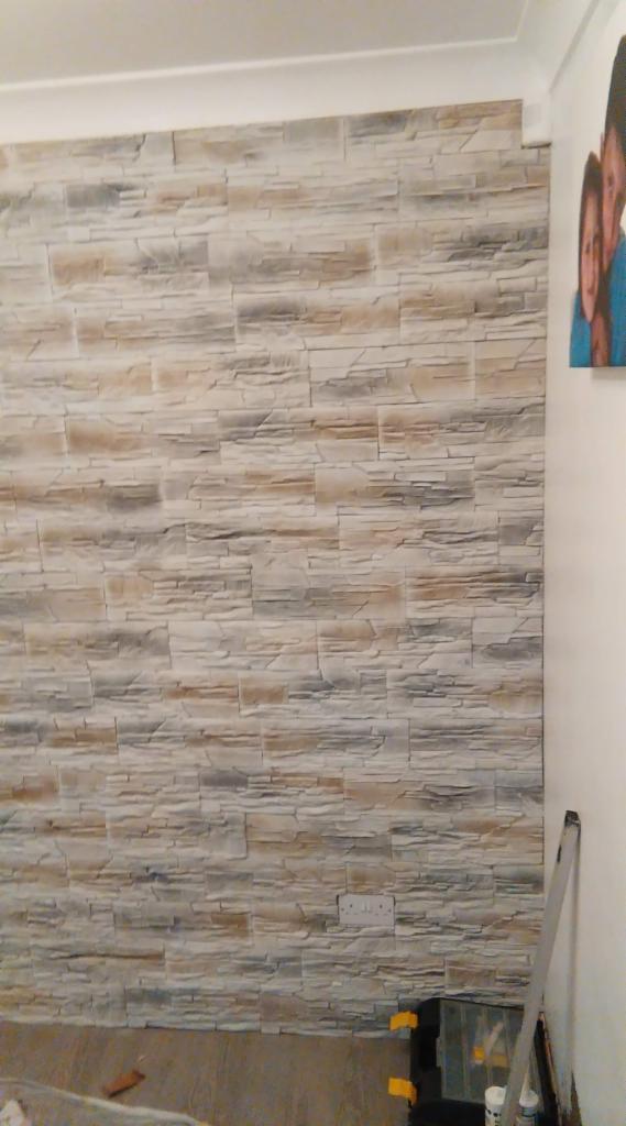 Ściana wykończona panelami imitującymi kamień, widoczna skrzynka z narzędziami i drabina, w tle obraz z wizerunkiem ludzi.