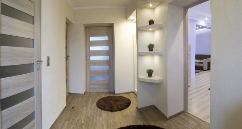 Jasne wnętrze przedpokoju z drewnopodobną podłogą, dwiema parami drzwi, w tym jedne z przeszkleniami, oraz wnęką z trzema półkami oświetlonymi punktowo, na których stoją małe doniczki z roślinami.