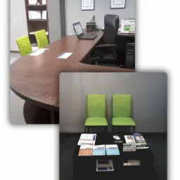 Biuro z dużym drewnianym stołem, zielonymi krzesłami, laptopem i dokumentami w ramkach na ścianie, dodatkowo stolik kawowy z materiałami informacyjnymi.