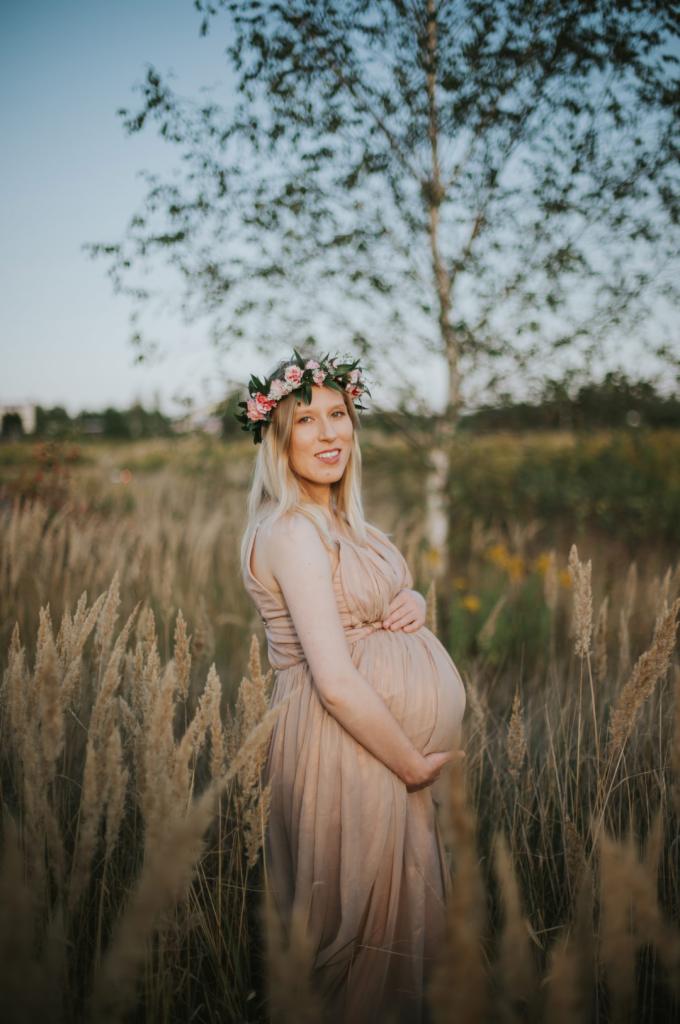 Kobieta w ciąży, w beżowej sukience i wianku z różowych kwiatów, pozuje w wysokiej trawie na tle drzewa brzozy.
