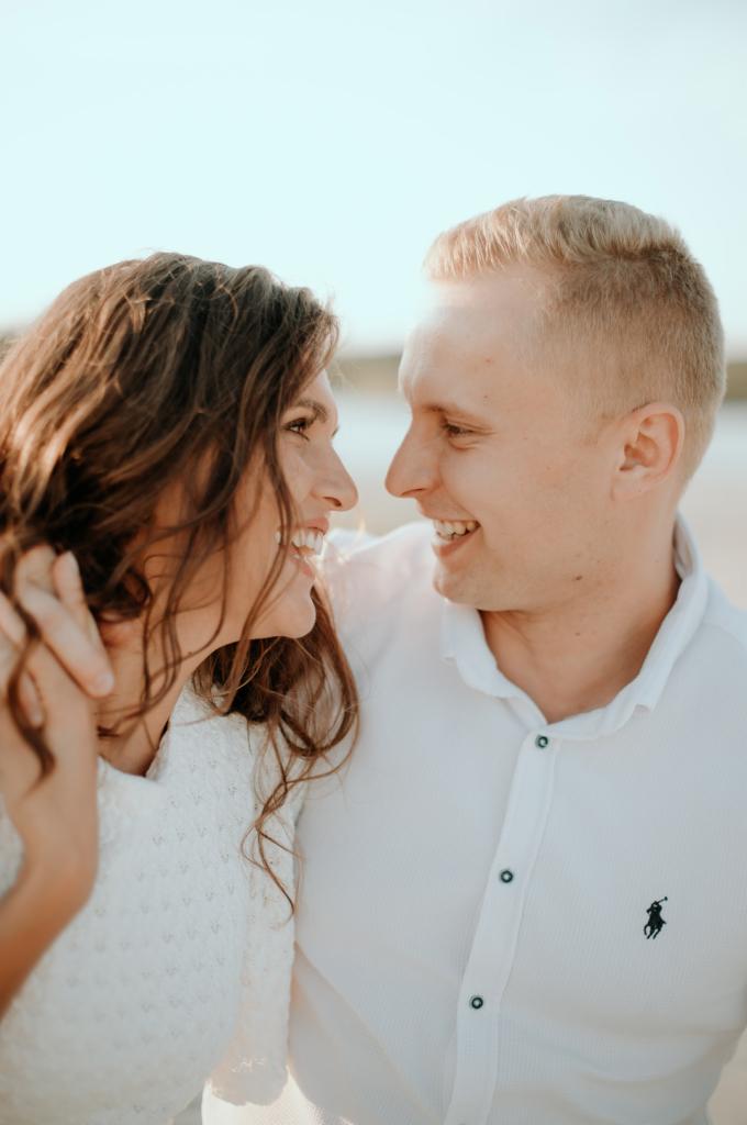 Uśmiechnięta para w białych ubraniach patrzy na siebie na tle jasnego nieba, z widocznym logo Polo na koszuli mężczyzny.