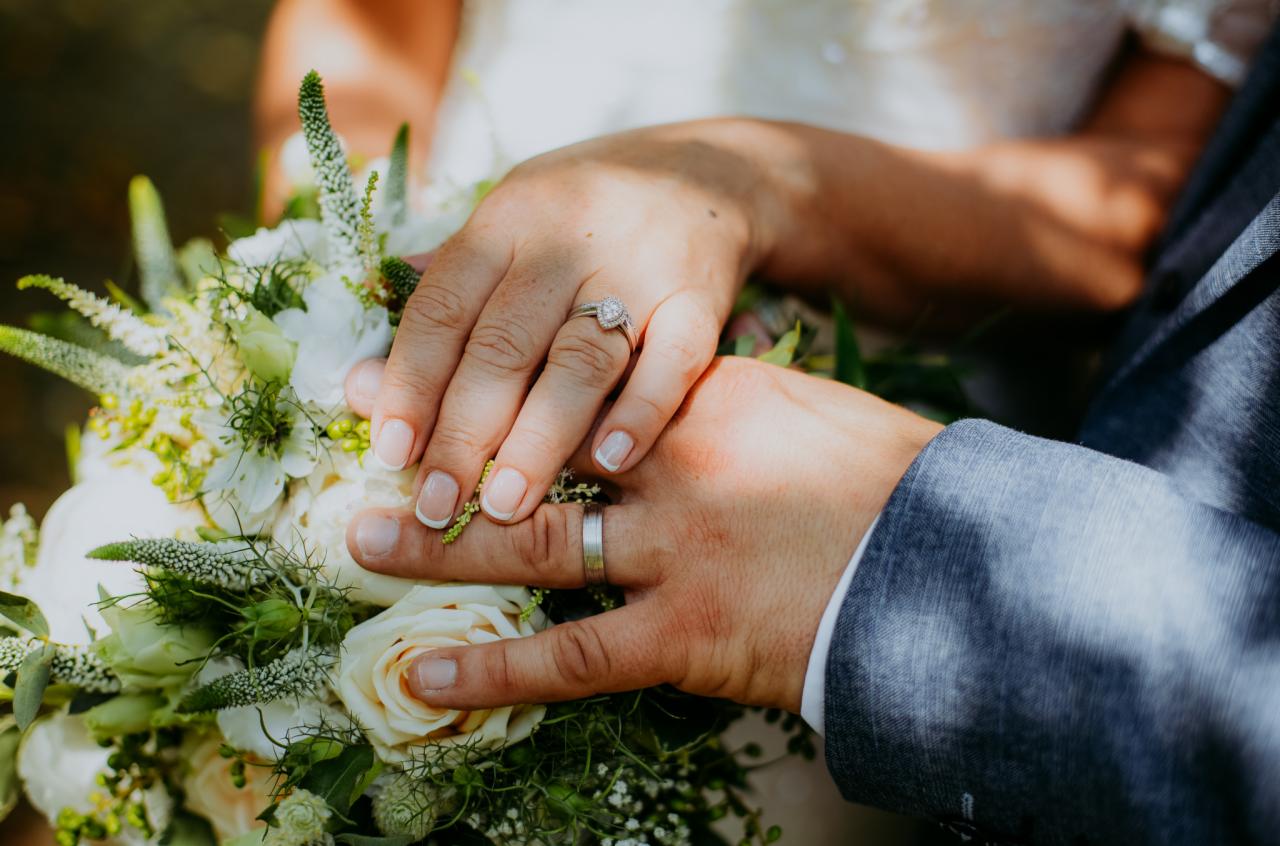 Dłonie pary młodej z obrączkami na tle bukietu ślubnego w odcieniach bieli i zieleni, pierścionek zaręczynowy z brylantem na palcu serdecznym.