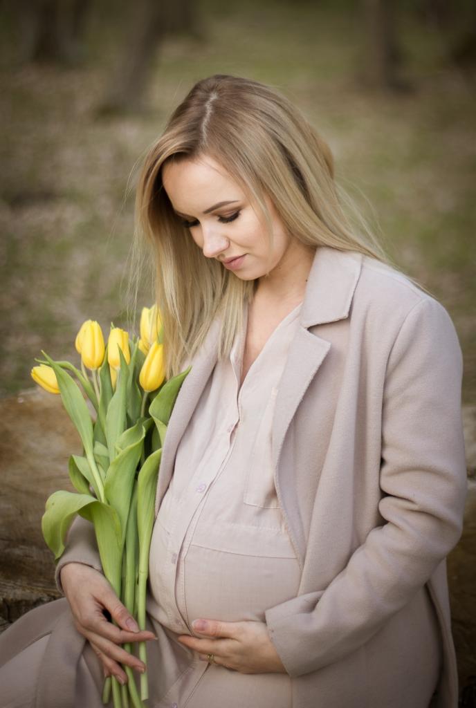 Kobieta w ciąży w beżowym płaszczu trzyma bukiet żółtych tulipanów, siedząc na pniu drzewa na zewnątrz.