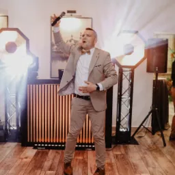 Mężczyzna w beżowym garniturze i muszce śpiewa do mikrofonu na tle sceny z oświetleniem punktowym podczas imprezy.