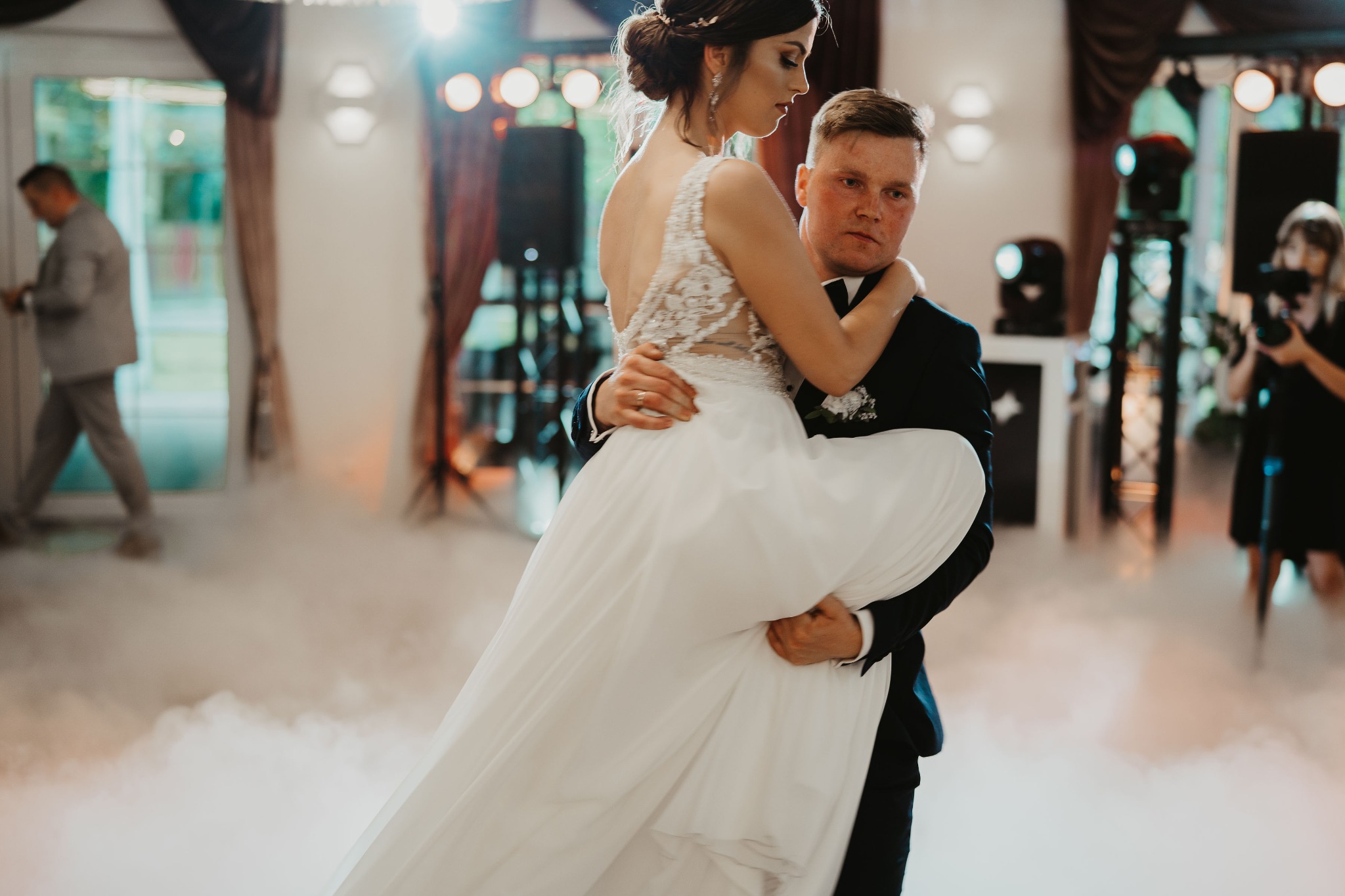 Pan młody trzyma pannę młodą w sukni ślubnej na rękach podczas pierwszego tańca w dymie scenicznym, w tle oświetlenie i fotograf.