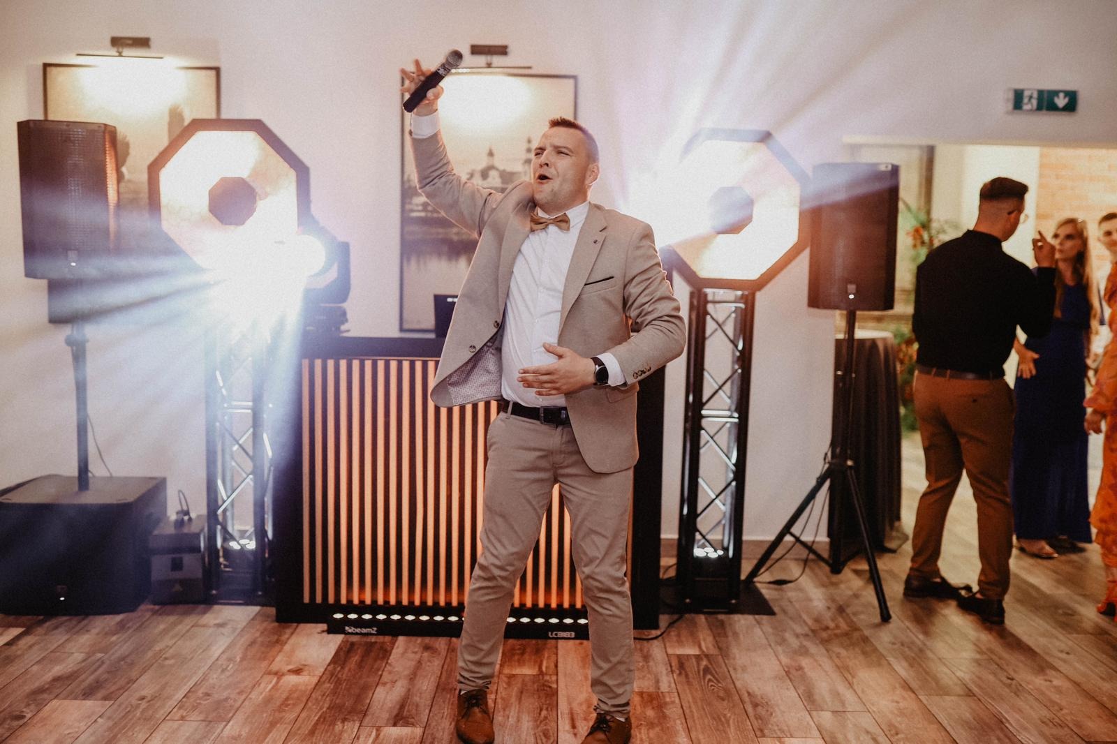 Mężczyzna w beżowym garniturze i muszce śpiewa do mikrofonu na tle sceny z oświetleniem punktowym podczas imprezy.