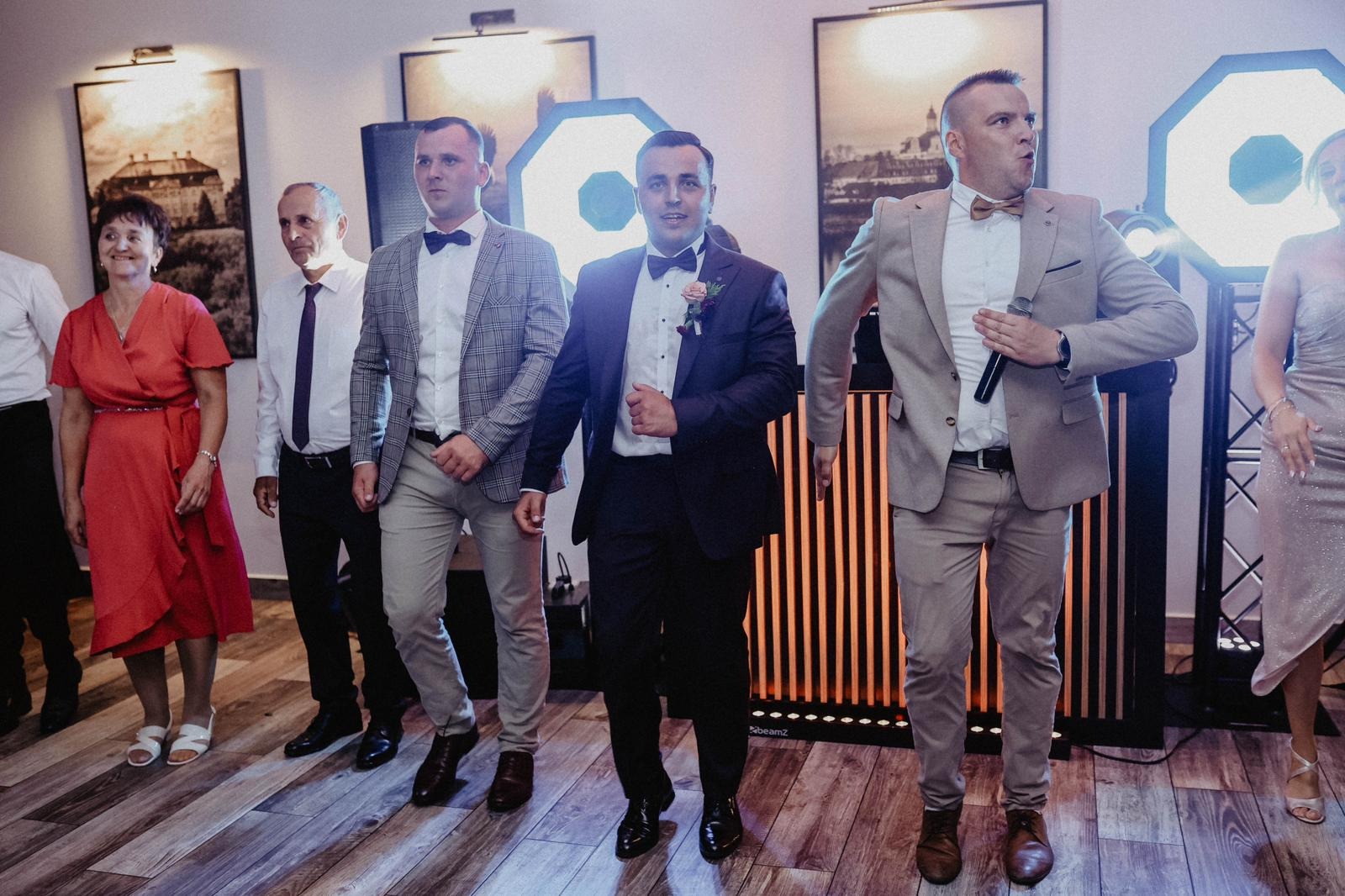 Zespół muzyczny w trakcie występu na weselu, wokalista z mikrofonem w dłoni i dwaj mężczyźni w garniturach tańczący na parkiecie, oświetlenie sceniczne w tle.