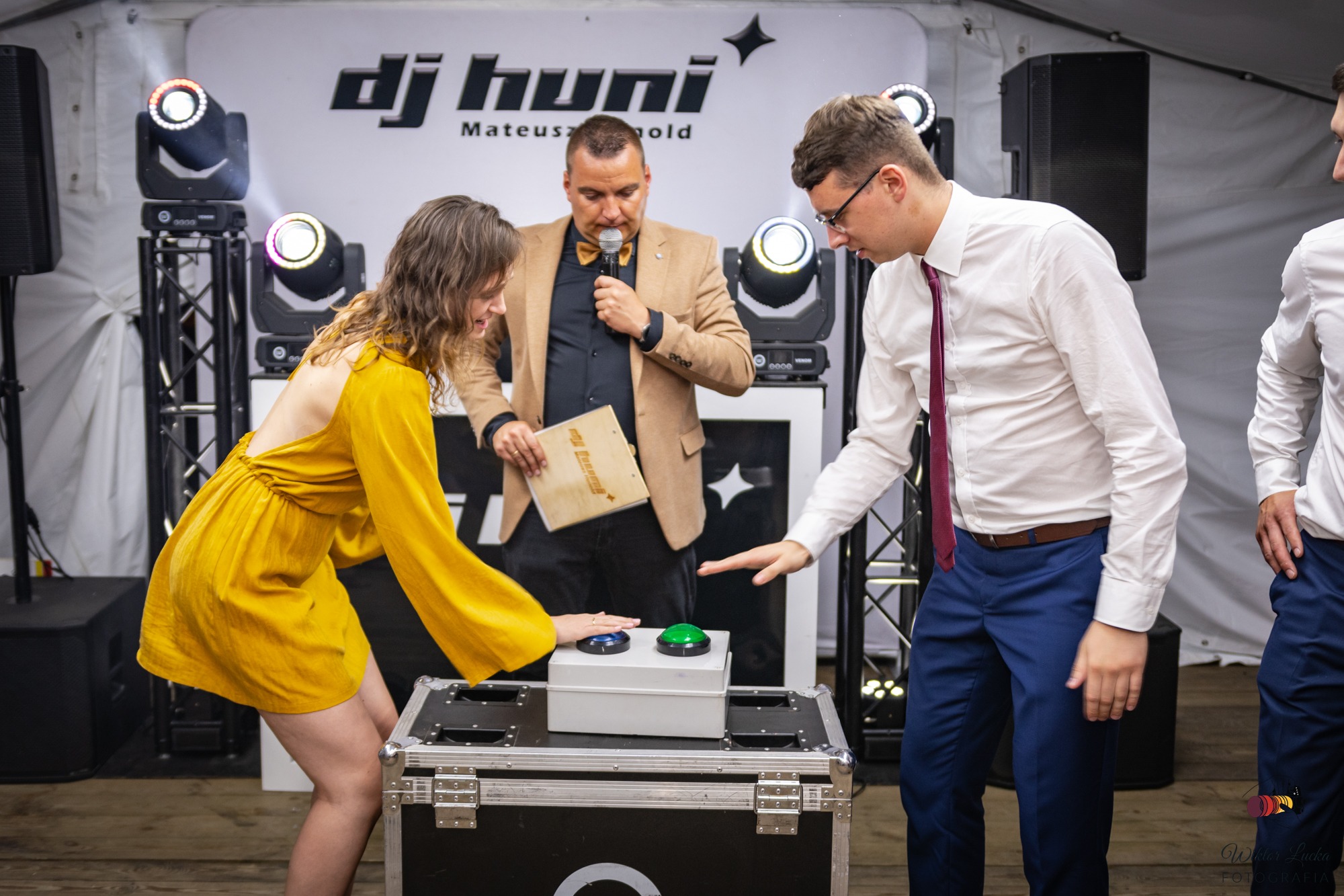 Prezenter w muszce prowadzi konkurs z dwoma uczestnikami naciskającymi przyciski na scenie z oświetleniem estradowym i logo DJ Huni.