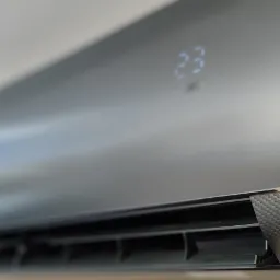 Zamontowany na ścianie, nowoczesny klimatyzator w kolorze grafitowym z wyświetlaczem LED pokazującym temperaturę 23 stopnie Celsjusza. Widoczna faktura imitująca włókno węglowe na dolnej krawędzi.