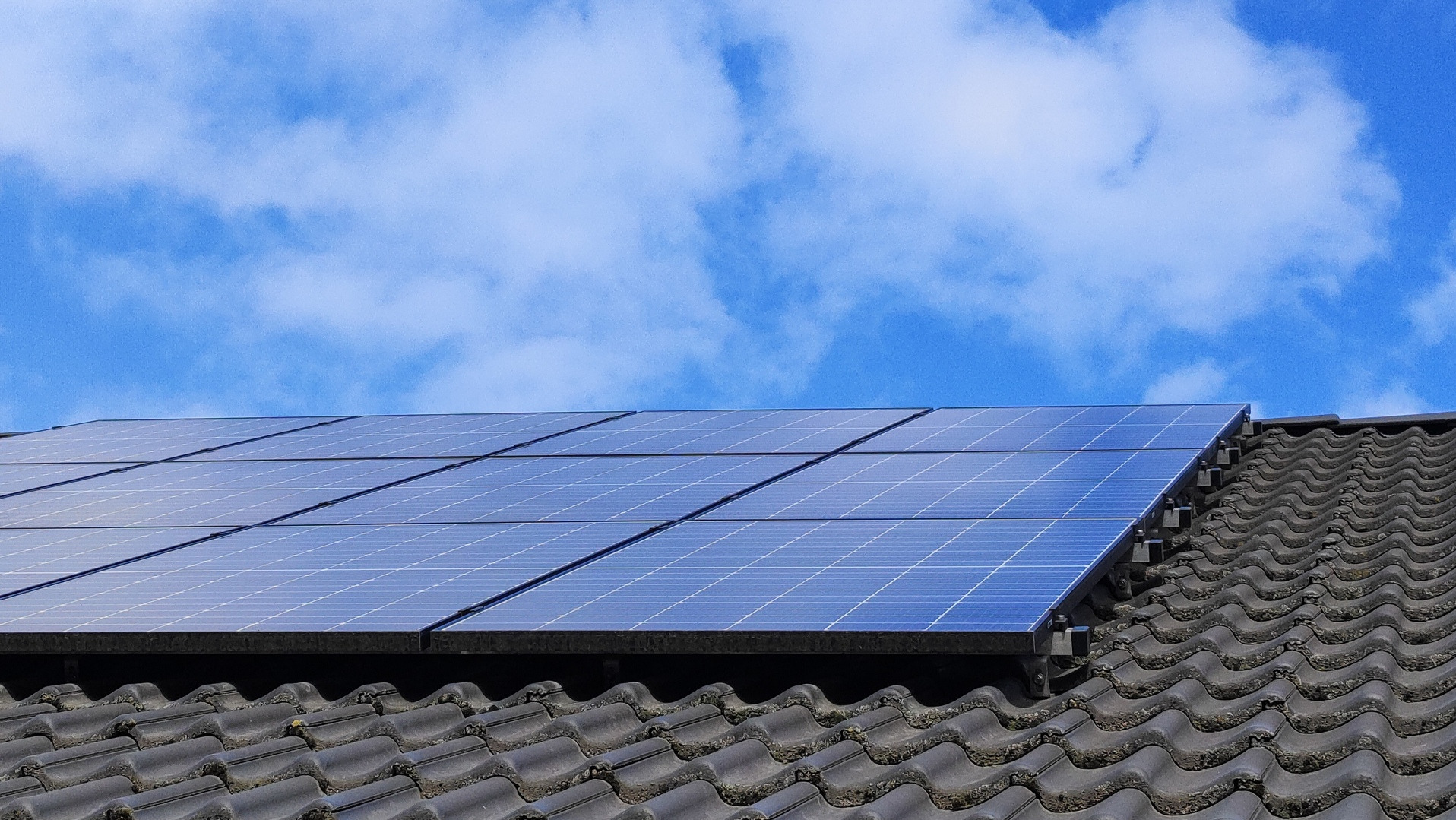 Panele słoneczne na ciemnoszarym dachu z dachówki ceramicznej, błękitne niebo z białymi obłokami w tle.