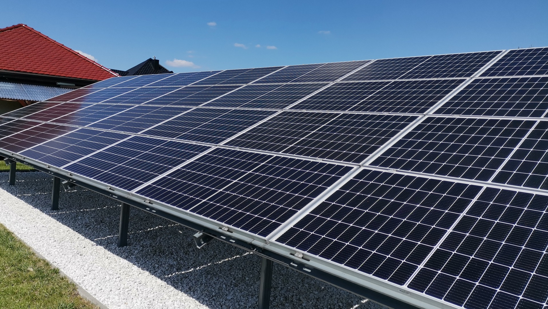 Instalacja paneli słonecznych na niskim stelażu, widok z boku, z czerwonym dachem budynku w tle i białym żwirem pod panelami, na tle błękitnego nieba.