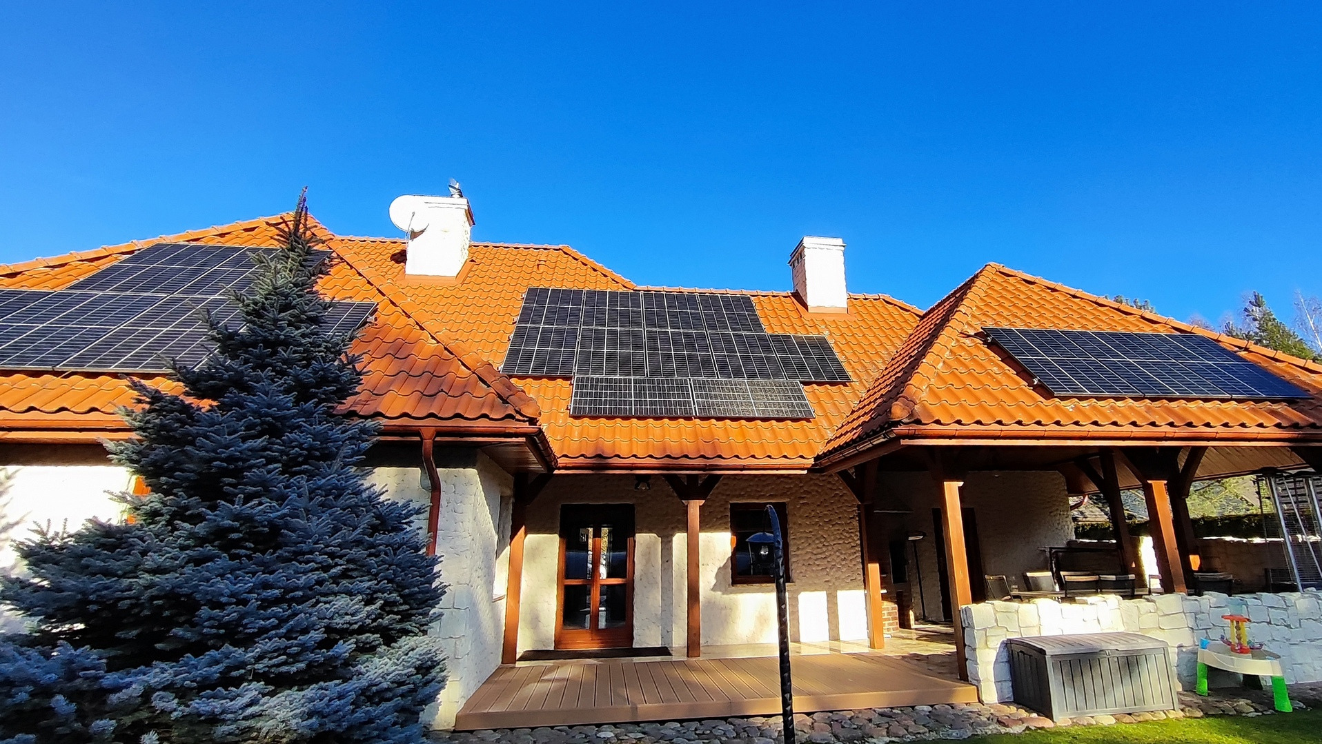 Dom z czerwoną dachówką i panelami fotowoltaicznymi na dachu, widok z poziomu ogrodu, z dużym świerkiem po lewej stronie.