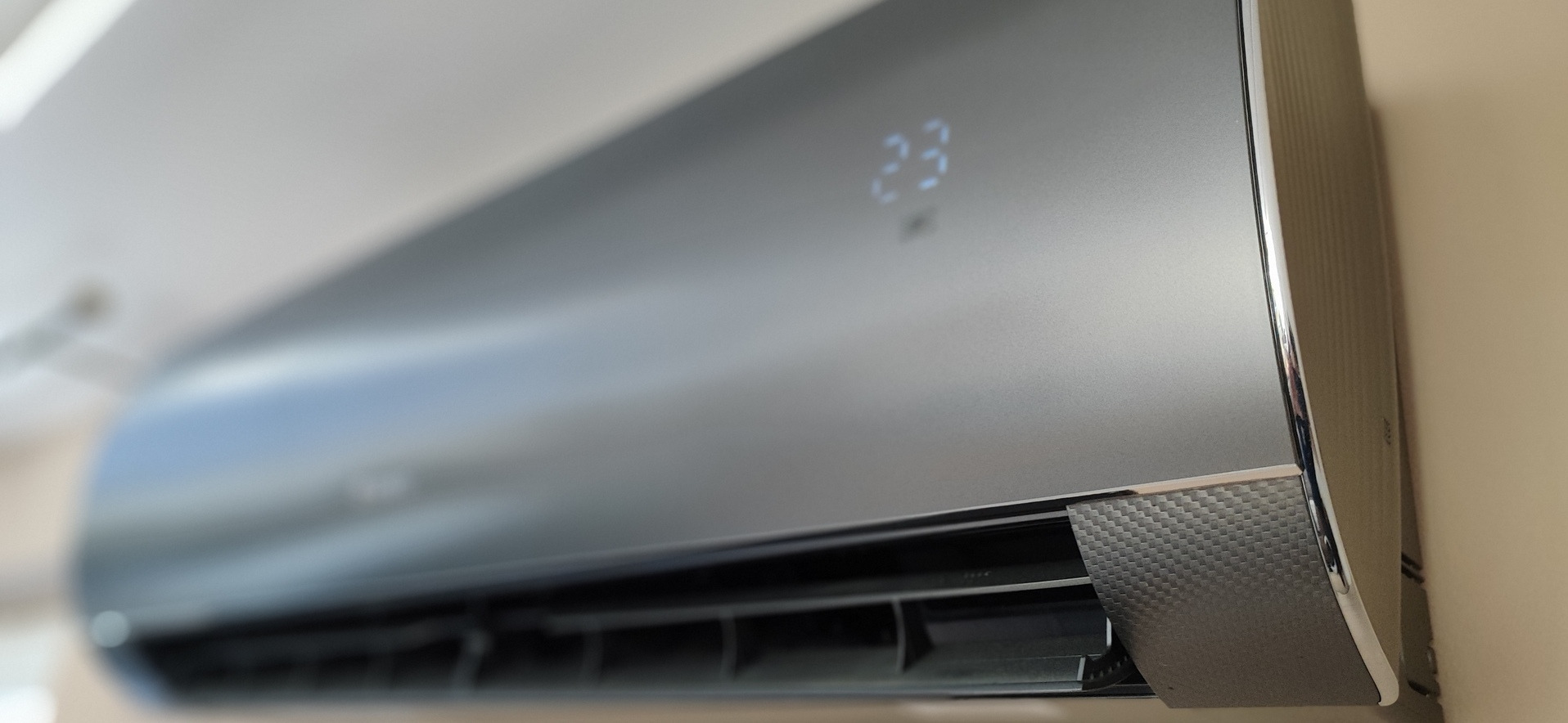 Zamontowany na ścianie, nowoczesny klimatyzator w kolorze grafitowym z wyświetlaczem LED pokazującym temperaturę 23 stopnie Celsjusza. Widoczna faktura imitująca włókno węglowe na dolnej krawędzi.