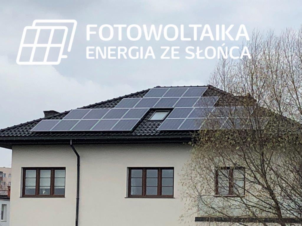 Dom jednorodzinny z panelami fotowoltaicznymi na dachu, widok z poziomu ulicy, na pierwszym planie gałęzie drzewa bez liści, w lewym górnym rogu logo firmy z napisem Fotowoltaika Energia ze Słońca.
